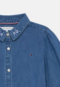 Τζιν πουκάμισο με κουμπιά μπροστά, λουλούδια κεντήματα στον γιακά, και μια μικρή ετικέτα λογοτύπου σε κόκκινο, λευκό και μπλε στο στήθος.