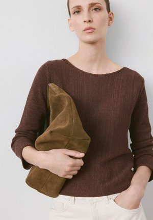 Femme portant un pull en tricot marron et un pantalon blanc, tenant une grande pochette souple marron sur un fond clair.