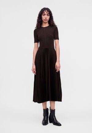 Femme aux cheveux bouclés portant une robe noire cintrée à manches courtes avec une jupe midi plissée et des bottines noires, debout sur un fond uni.