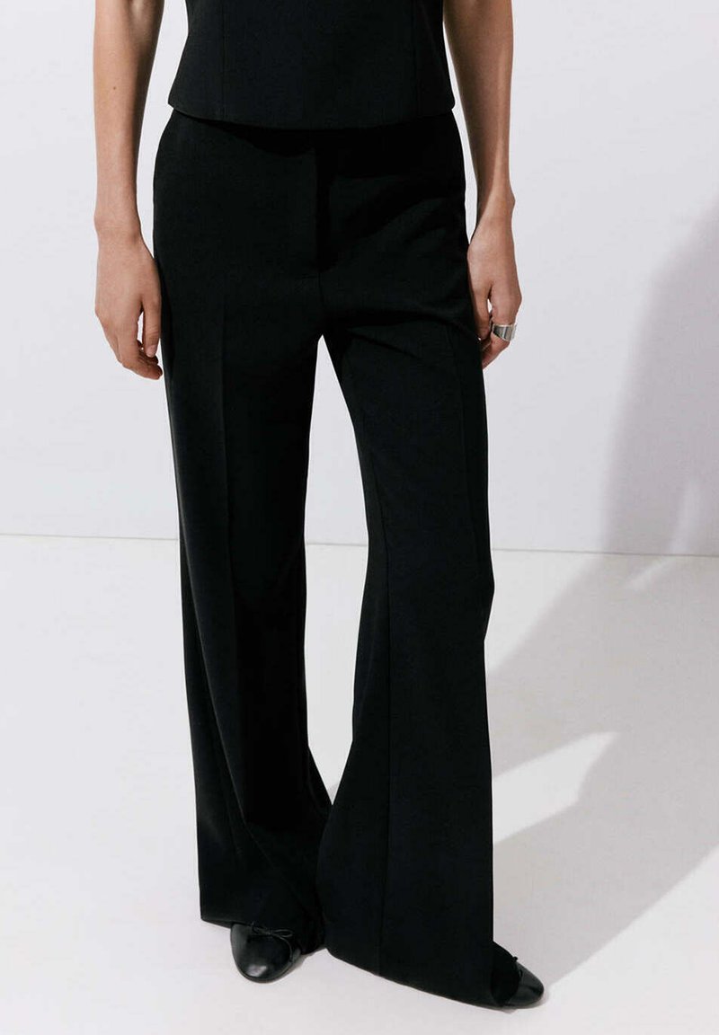 Personne portant un pantalon noir à jambes larges, des chaussures noires pointues et un haut noir, debout sur un fond blanc uni.