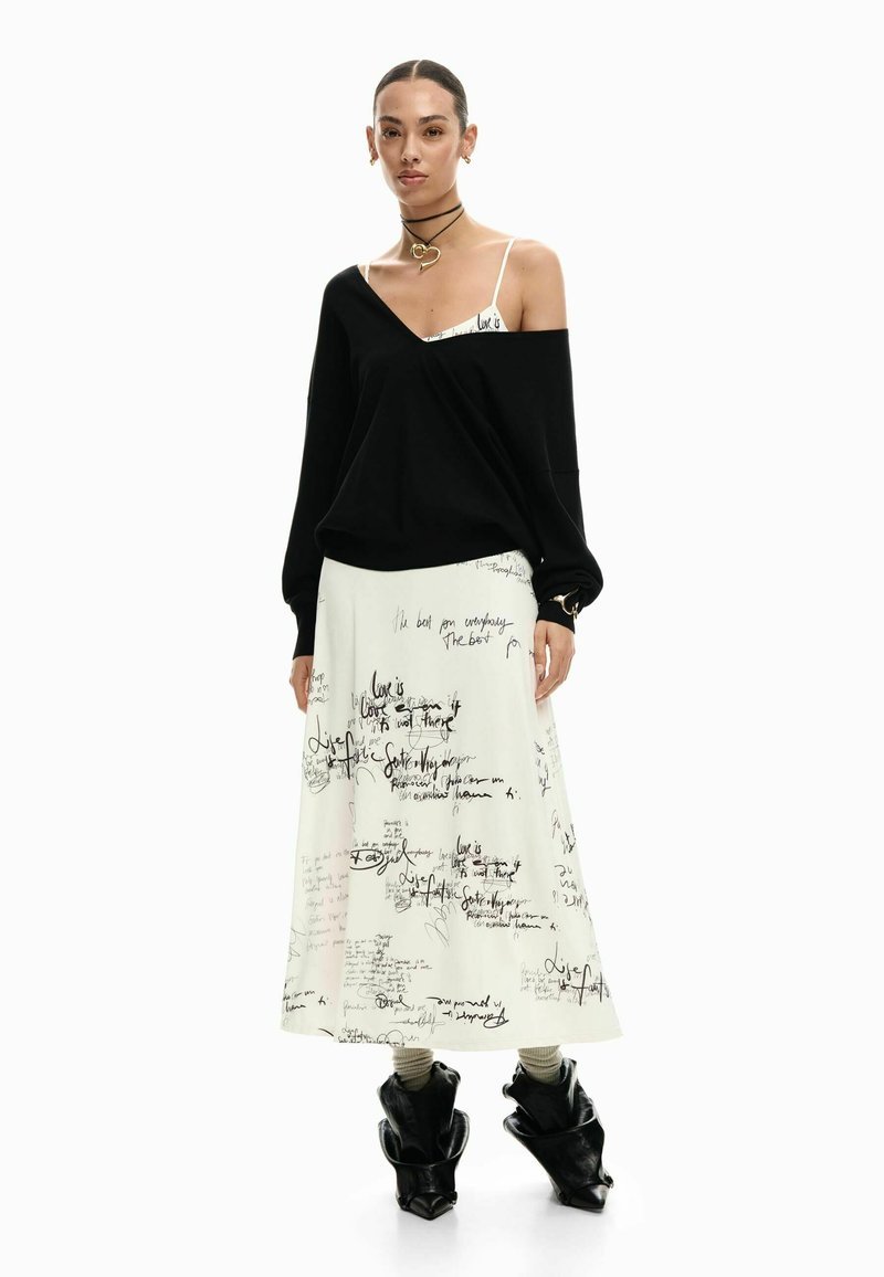 Fekete, túlméretezett pulóver V-nyakkivágással, fehér szoknyával, amelyen kézzel írt fekete szöveg található, valamint fekete, szoborszerű bokacsizmával párosítva.