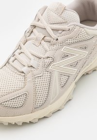 Beige athletic shoe with mesh upper, lacing system, and rubber sole featuring textured accents and logo. Smooth, breathable texture.