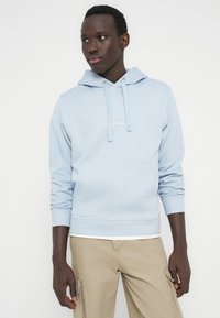 Mężczyzna w jasnoniebieskiej bluzie z kapturem i beżowych spodniach typu cargo, stojący na tle jednolitego białego tła, patrzący nieco w lewo.