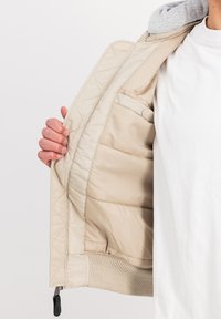 Beige quilted bomber jacket with a gray hood, featuring a ribbed hem and an inner pocket, displayed open to show the lining and texture.