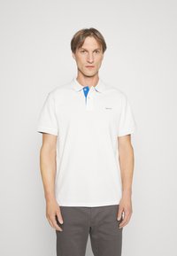 Weißes Poloshirt mit einem blauen Button-Down-Kragen, gesticktem Logo und kurzen Ärmeln, kombiniert mit grauen Hosen. Glatte Stofftextur.