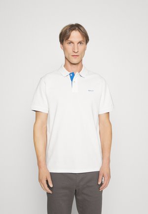 Hvit polo skjorte med blå nedbrettbar krage, brodert logo og kort erm, kombinert med grå bukser. Glatt stofftekstur.