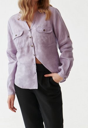 Femme portant une chemise lilas claire à boutons avec deux poches poitrine et un pantalon noir, une main dans la poche, sur un fond uni.