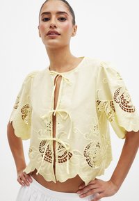 Rumena vezenina bluzica s kratkimi napihnjenimi rokavi, sprednjim vezanjem, valovitim robom in odprtimi čipkastimi detajli. Bombažni material, lahek tekstur.
