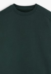 Sudadera verde oscuro con cuello redondo y tejido texturizado. Presenta un sutil logo en la esquina inferior. Superficie suave y diseño ajustado.