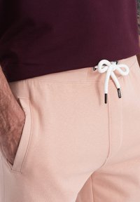 Pantalons de survêtement rose clair en tissu doux, dotés d'une taille élastique avec cordon blanc et de poches avant. Design simple et fonctionnel.