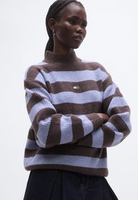 Csíkos pulóver kötött anyagból; váltakozó világoskék és barna csíkokkal, magas nyakkal és egy kis logóval a mellkasán.