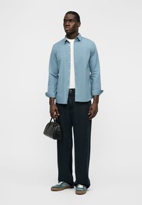Jeune homme portant une chemise bleu boutonnée, un t-shirt blanc, un jean noir à jambes larges, des baskets vertes, tenant un petit sac à main noir.