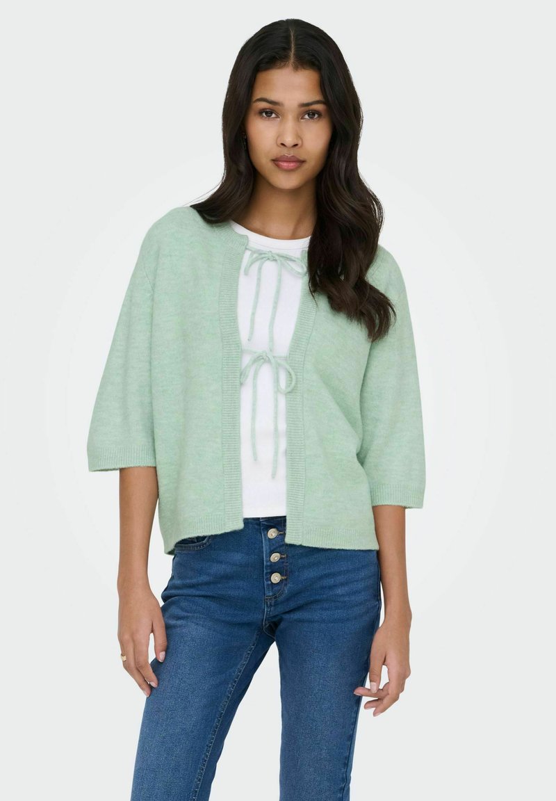 Cardigan vert clair à manches trois-quarts, ouvert sur le devant avec une finition texturée. Porté sur un haut blanc avec un détail noué à l'avant. Jean bleu visible.