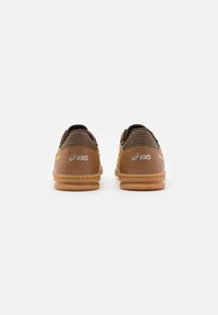 Chaussures de sport brunes avec une finition en cuir lisse, semelles en caoutchouc et accents texturés ; dotées d'une marque jaune et d'un détail cousu au talon.