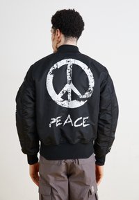 Black bomber jacket featuring a large white peace symbol and the word "PEACE" printed on the back. Ribbed cuffs and hem. Smooth texture.