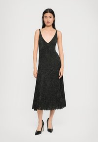 Mulher vestindo um vestido midi plissado preto sem mangas com um decote em V profundo e sapatos de salto alto pretos, posicionada contra um fundo simples.