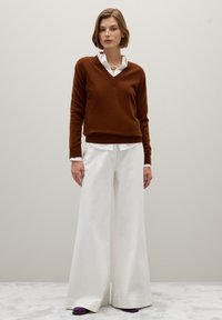 Un pull brun en V porté sur une chemise blanche à col, associé à un pantalon blanc à larges jambes et des chaussures violettes, le tout sur un fond uni.