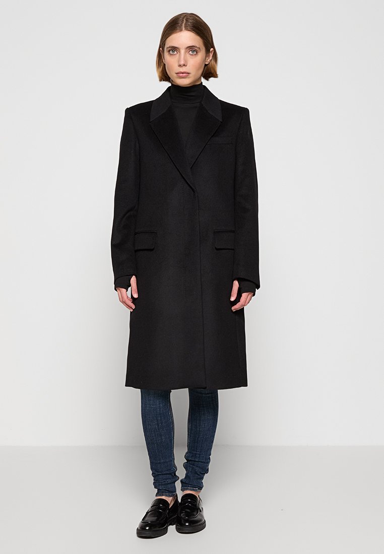 Manteau en laine noir avec un col revers, deux poches avant et une silhouette droite. Porté sur un col roulé noir et un jean bleu.