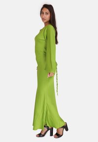 Lang grøn satin kjole med lange ærmer, sideslids og flettet detalje; båret med sorte åbne hæle. Glat tekstur og tætsiddende design.