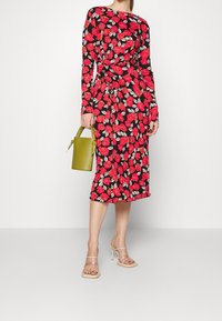 Kvinna i en svart midiklänning med röd blommig mönster, nude högklackade sandaler och håller i en liten olivgrön handväska.