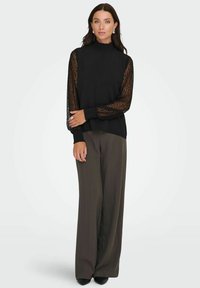 Zwarte top met hoge hals en lange doorschijnende kanten mouwen, gecombineerd met wijde donkergekleurde broek. Gladde stoftextuur, gelaagd ontwerp met details.