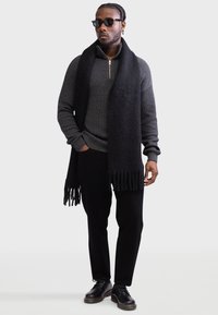 Black knit sweater with a zippered neck, paired with black trousers and a long black scarf featuring fringed edges, complemented by black shoes.