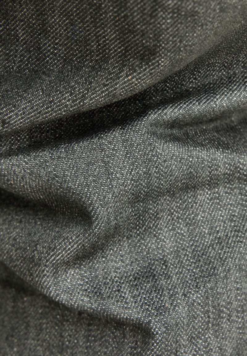 Close-up van textuur van donkergrijs geweven stof met zichtbare diagonale weving en zachte plooien die schaduwen en diepte creëren.