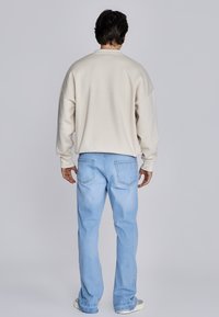 Beige sweatshirt met verlaagde schouders, gecombineerd met lichtblauwe spijkerbroek. Eenvoudig ontwerp; losse pasvorm, zichtbare stiksels en achterzakken.