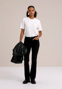 Bela kratka majica s kratkimi rokavi, visoke črne jeans hlače, črna usnjena jakna obešena na roki, črne čevlje, model stoji s roko na boku.