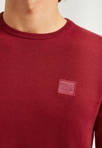 Burgundowa sweter z dzianiny z okrągłym dekoltem i teksturowanym wykończeniem. Posiada kwadratową, wytłaczaną łatkę z logo "POLO CLUB" na piersi.