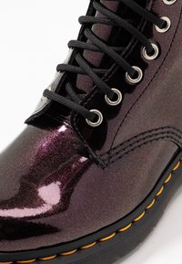 Botín con cordones en negro brillante con efecto purpurina púrpura. Presenta ojales metálicos, cordones negros y costuras amarillas a lo largo del borde de la suela.