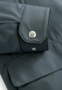 Détail d'une veste de pluie gris foncé avec un poignet boutonné équipé de ferrures argentées et une texture de tissu lisse et résistant à l'eau.