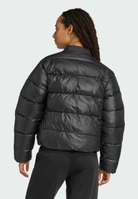 Osoba s dlhými, tmavými vlasmi je viditeľná zozadu, oblečená v nadýchanej čiernej bunde a čiernych nohaviciach na svetlošedom pozadí.