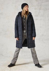 Abrigo acolchado largo azul marino con capucha, que presenta un diseño acolchado. Lleva puesto sobre una camisa negra de botones y jeans grises, combinados con botas negras.