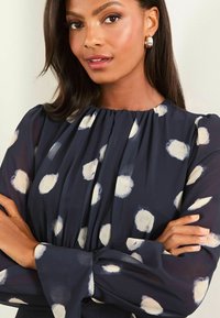 Bluză navy cu model de puncte albe, cu față pliată și mâneci lungi. Material transparent cu decolteu și manșete elasticizate.