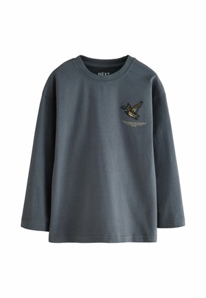 Ragazzo in piedi con le mani nelle tasche, indossa una maglia a maniche lunghe grigio scuro, pantaloni cargo mimetici marroni e scarpe da ginnastica bianche.
