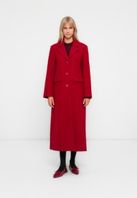 Red long coat with a notched collar, two front pockets, and button closure. Worn over a black top and paired with black tights and red shoes.