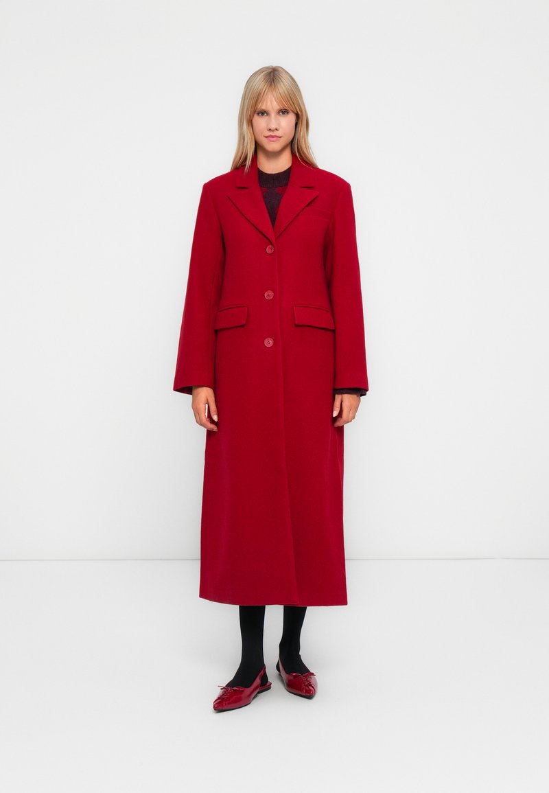 Red long coat with a notched collar, two front pockets, and button closure. Worn over a black top and paired with black tights and red shoes.