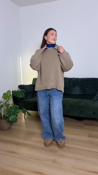 Ylimittainen beige neulepusero, sininen huivi, väljät vaaleansiniset farkut, vaaleanruskeat slip-on-kengät, seisoo puulattialla lähellä vihreää sohvaa ja kasvia.