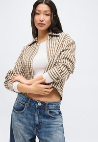 Νεαρή γυναίκα με μακριά σκούρα μαλλιά φοράει καφέ καρό μπουφάν πάνω από λευκό τοπ και μπλε τζιν, στέκεται με τα χέρια σταυρωμένα μπροστά σε λευκό φόντο.