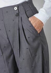 Sive plisirane hlače s teksturiranim materijalom, s patentnim zatvaračem, dva gumba i bočnim džepovima. Male uzorčaste dekoracije raspršene po cijelom modelu.