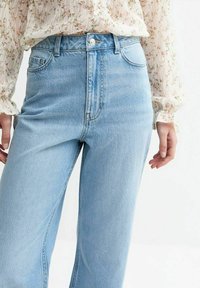 Osoba w jasnoniebieskich spodniach o wysokim stanie i białej bluzce z długim rękawem w kwiatowy wzór, stojąca na jednolitym tle.