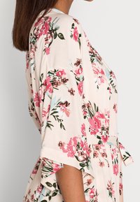 Kwiatowy kimono w lekkim materiale, z miękkim beżowym tłem i różowymi oraz zielonymi kwiatami, szerokimi rękawami i zawiązywanym w kroku paskiem.