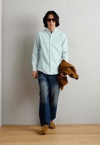 Light blue and white striped shirt, blue jeans, brown jacket held in hand, brown shoes, and sunglasses. Simple design, relaxed fit.