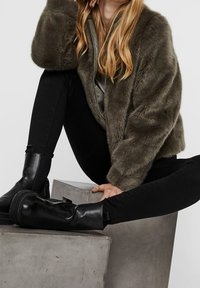 Person i en brun kunstpelsjakke, sorte skinny jeans og sorte læderstøvler, siddende på og ved siden af grå betonblokke.