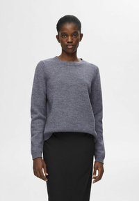 Grå strikket sweater med rund hals og lange ærmer. Har en afslappet pasform og ribbet manchetter, parret med en sort nederdel.