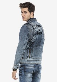 Denim jacket met metalen schouderaccenten, vervaagde blauwe was, ritszakken en geborduurde tekst op de rug. Verweerde textuur detail.