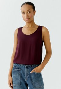 Burgundy sleeveless top with a scoop neckline, made of smooth fabric, paired with blue jeans featuring front pockets and a relaxed fit.