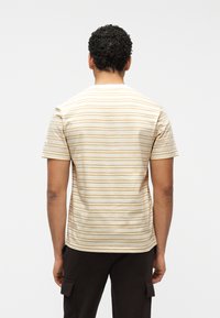 Άνδρας με σγουρά μαλλιά, ντυμένος με κοντομάνικο πουκάμισο σε μπεζ και άσπρες ρίγες και σκούρο παντελόνι, στέκεται με την πλάτη προς τα εμπρός σε απλό φόντο.