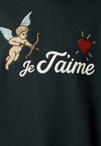 Chérubin brodé avec un arc et une flèche visant un cœur rouge, avec le texte en écriture blanche « je T'aime » sur fond de tissu sombre.